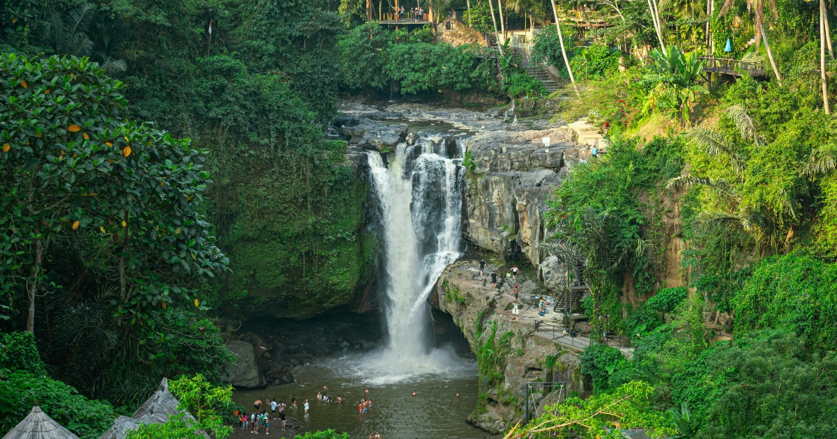 A breathtaking view of Tegenungan Waterfall near Ubud, Bali. The cascading waterfall flows down into a natural pool surrounded by lush green cliffs, while visitors swim and relax below. Dense jungle and tropical vegetation frame the scene, with pathways and viewing platforms built into the hillside.